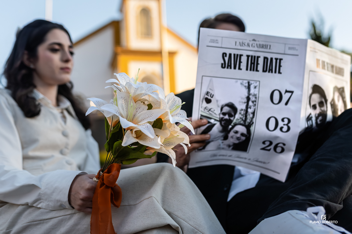 Laís segura um buquê de flores brancas e olha com carinho para Gabriel, que carrega um jornal antigo, sentados na praça em Santo Antônio do Pinhal