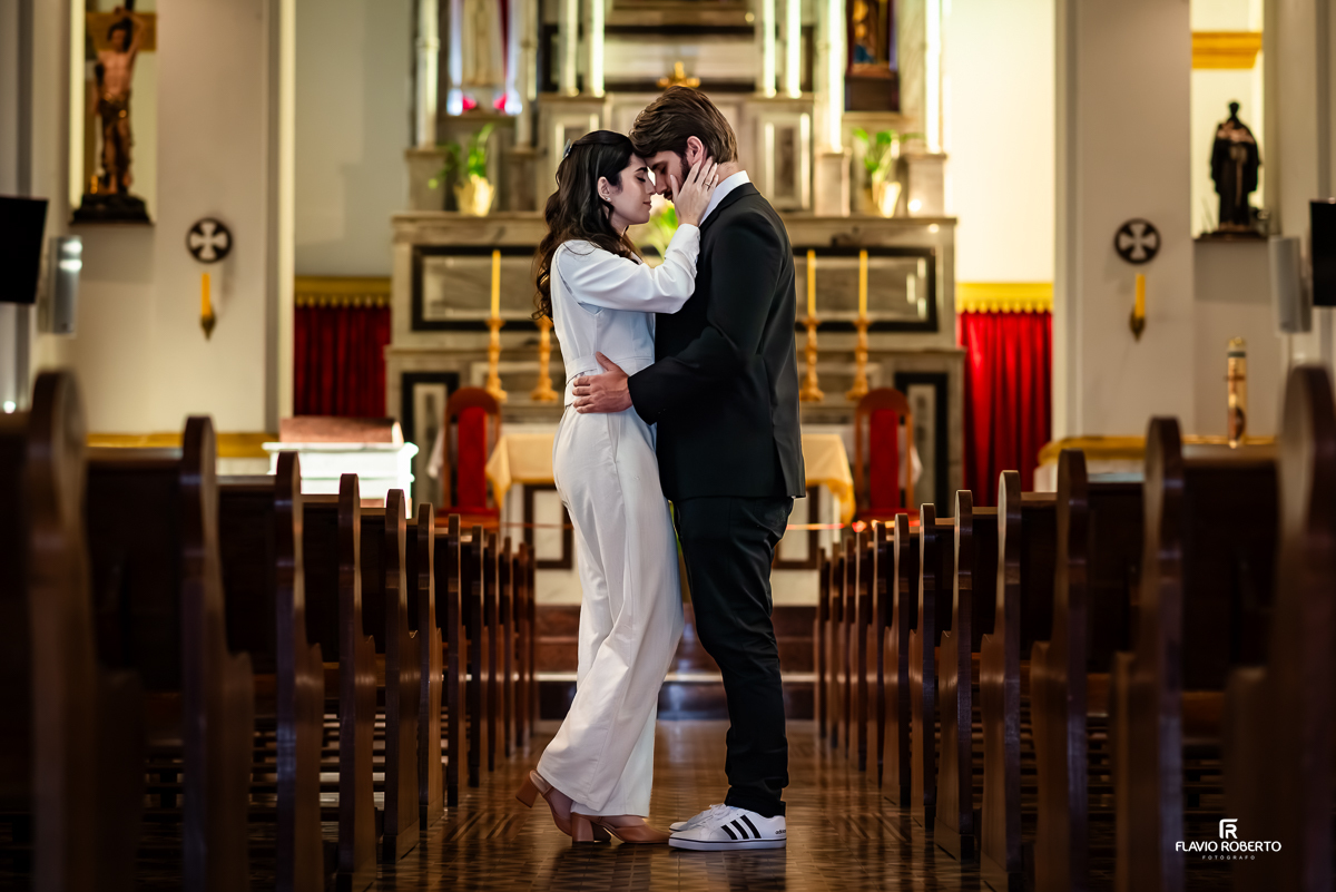 Casal Laís e Gabriel se abraçam com carinho no corredor central de uma igreja, vestidos de forma elegante e minimalista, com fundo do altar suavemente iluminado.