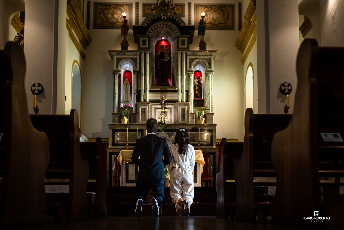 Laís e Gabriel ajoelhados lado a lado no altar da igreja, em um momento de oração íntimo e simbólico, diante de um altar clássico com imagens religiosas