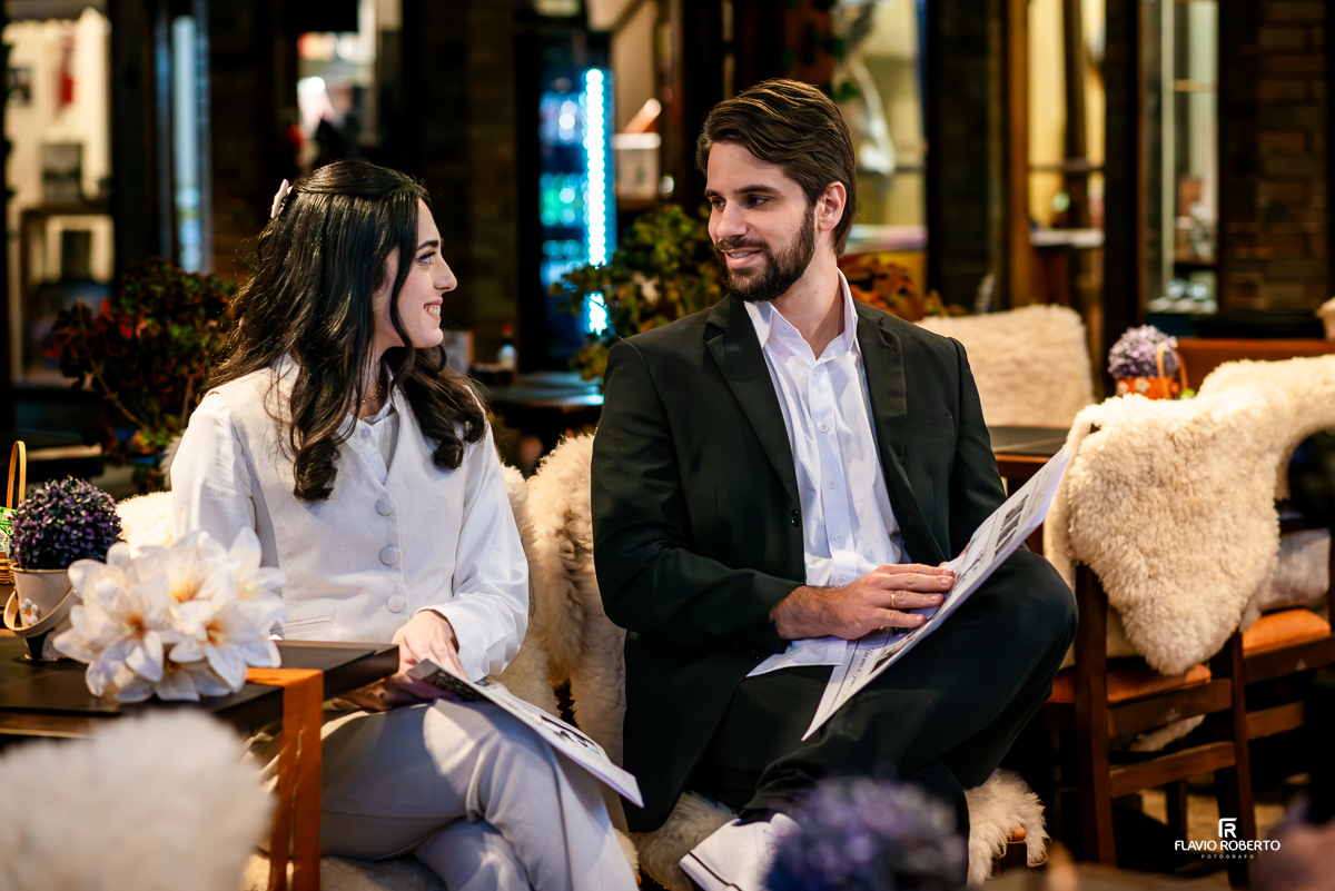 Laís e Gabriel sentados, sorrindo e segurando jornais personalizados com a data do casamento e fotos do casal, em ambiente decorado com luzes e flores