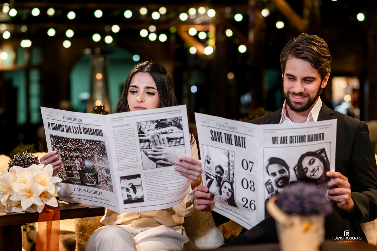 Laís e Gabriel sentados, sorrindo e segurando jornais personalizados com a data do casamento e fotos do casal, em ambiente decorado com luzes e flores