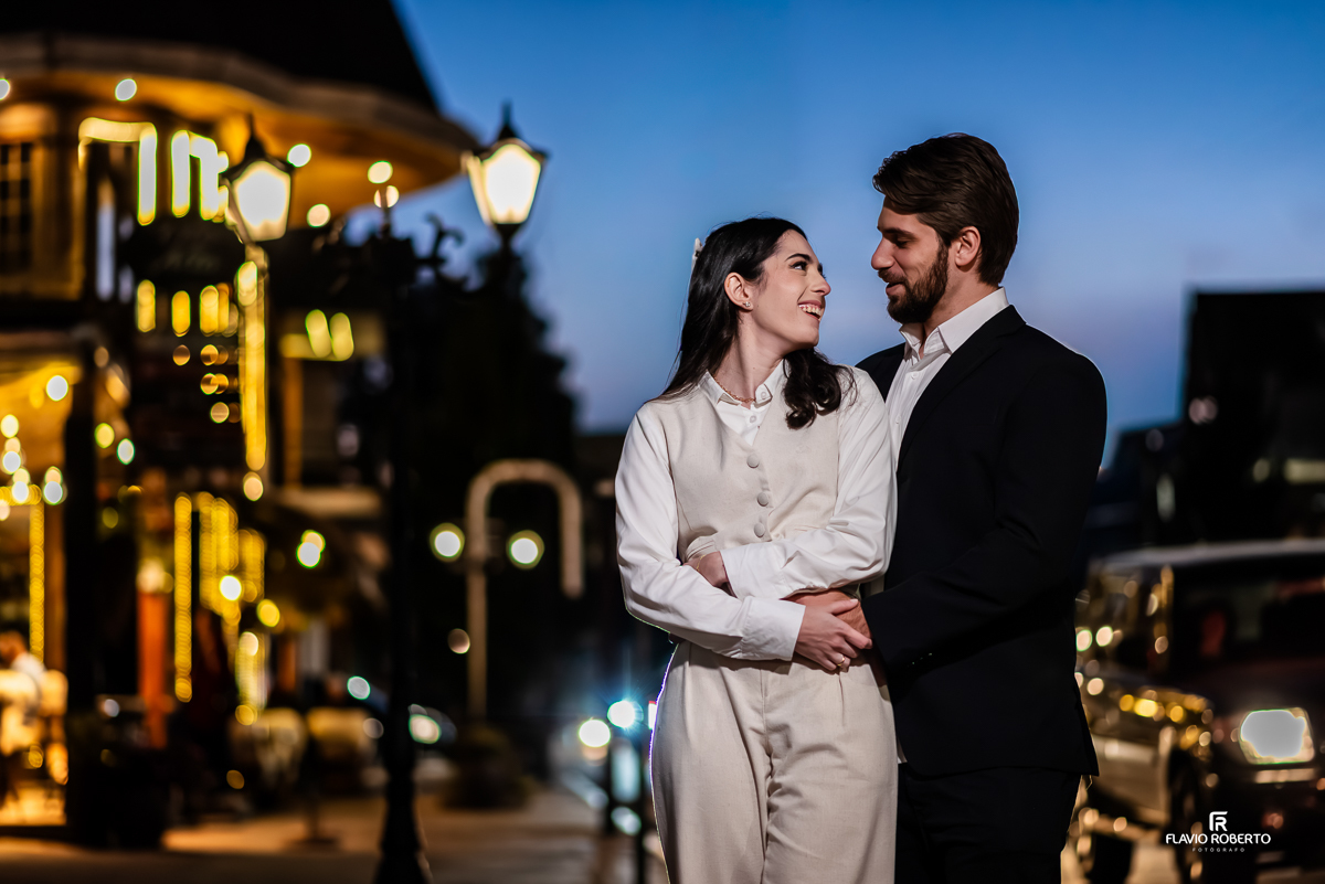 Laís e Gabriel caminham abraçados pelas ruas iluminadas de Santo Antônio do Pinhal ao anoitecer, trocando olhares sorridentes em um clima romântico.
