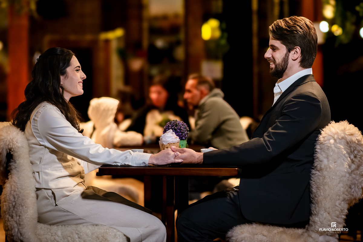 Laís e Gabriel sentados à mesa de um café, sorrindo um para o outro enquanto seguram as mãos sobre a mesa decorada com flores e sobremesa.