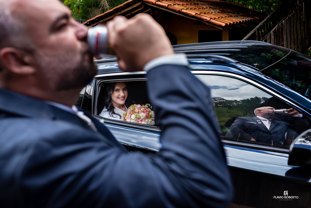 Noivo brindando após o casamento enquanto a noiva sorri dentro do carro em Campos de Cunha