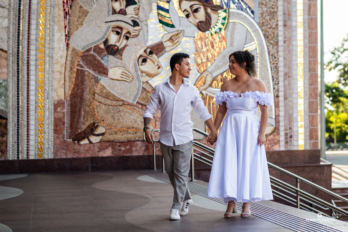 Casal caminhando em frente a painel religioso no Santuário Nacional de Aparecida durante ensaio pré wedding