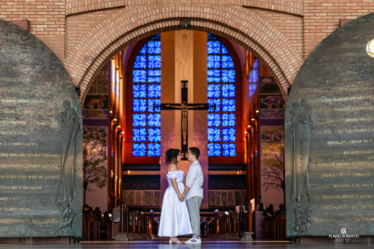 Casal posicionado no interior do Santuário Nacional de Aparecida com cruz ao fundo durante ensaio pré wedding
