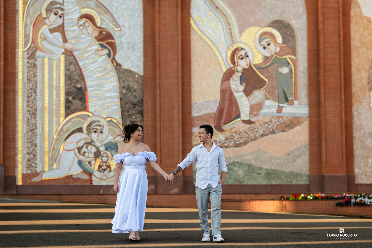 Casal caminhando em ensaio pré wedding no Santuário Nacional de Aparecida com painel religioso ao fundo