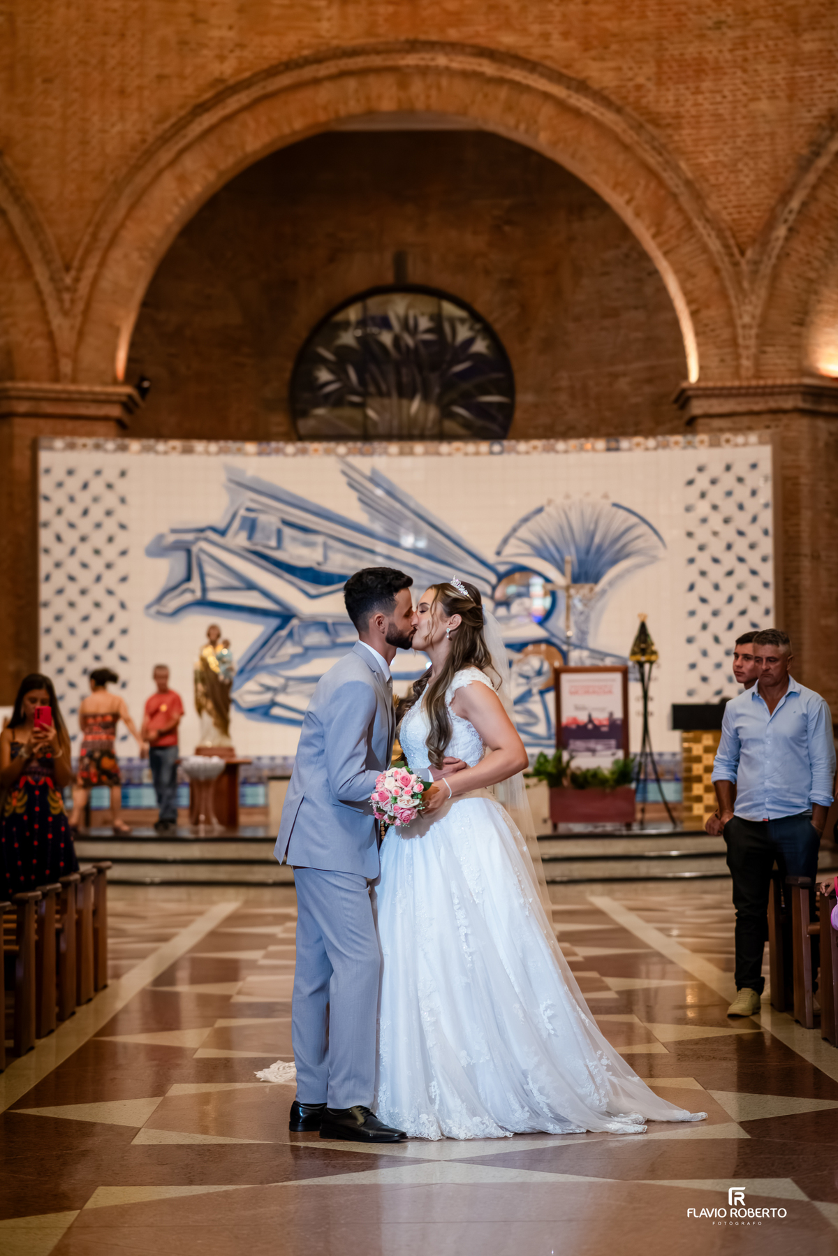 Beijo dos noivos durante casamento no Santuário Nacional de Aparecida com convidados ao fundo