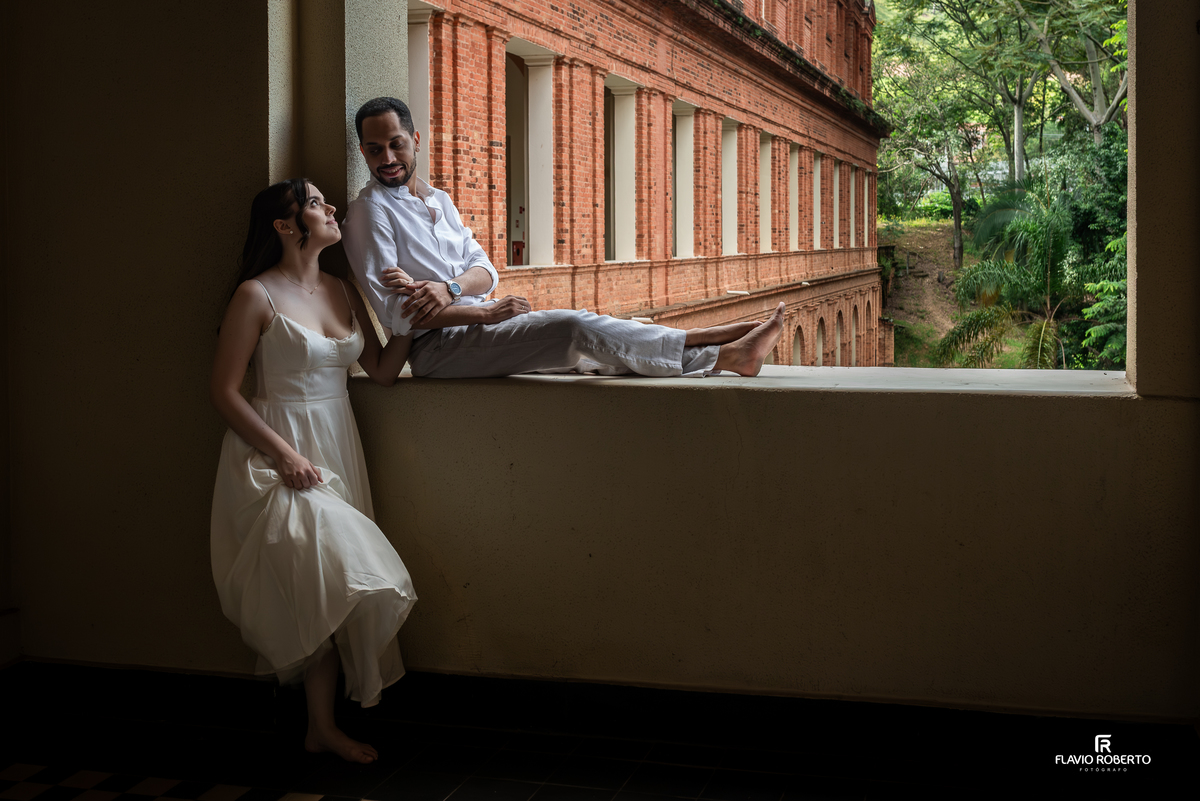 Casal em janela do Seminário Bom Jesus em Aparecida com fachada histórica de tijolos ao fundo