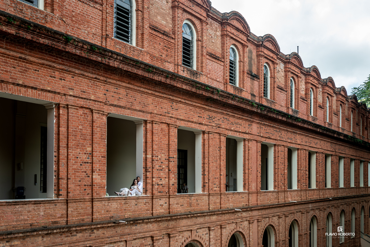 Casal em janela do Seminário Bom Jesus em Aparecida com fachada histórica de tijolos ao fundo