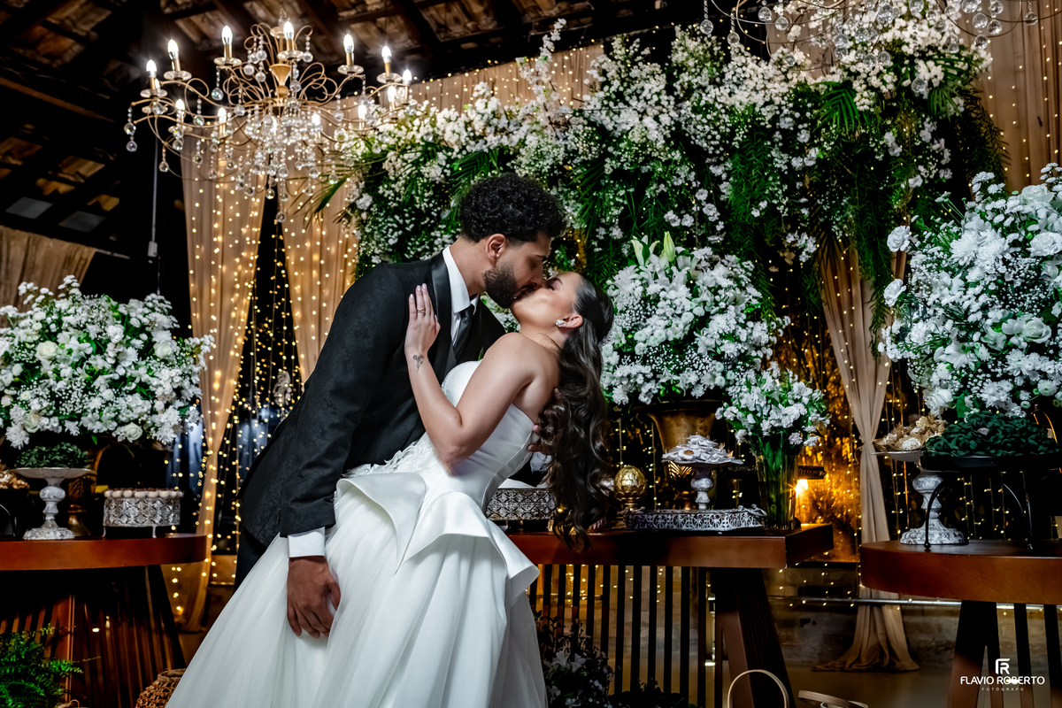 Casal trocando um beijo apaixonado diante da decoração iluminada e floral da recepção de casamento no Sítio Tangaroa em Taubaté.