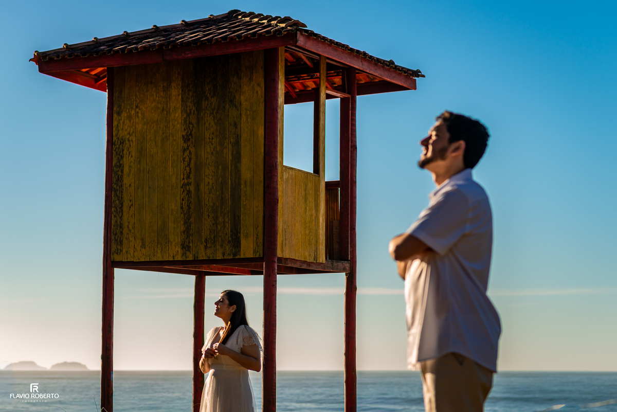 Casal durante ensaio pré wedding com a tradicional guarita da Praia Vermelha do Norte ao fundo, em Ubatuba.