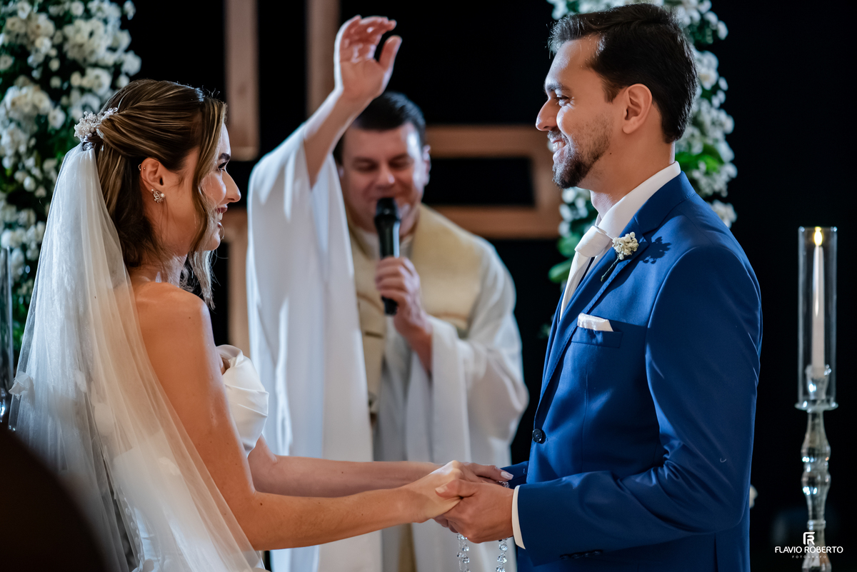 Noivos de mãos dadas recebem a bênção do sacerdote durante casamento católico no Recanto do Bosque.