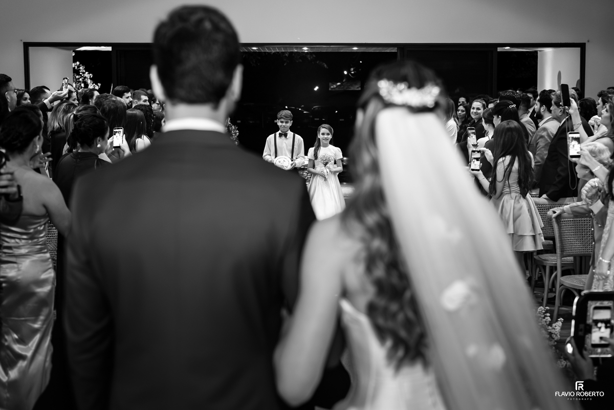 Noivos vistos de costas enquanto crianças entram pelo corredor da cerimônia de casamento em Guaratinguetá.