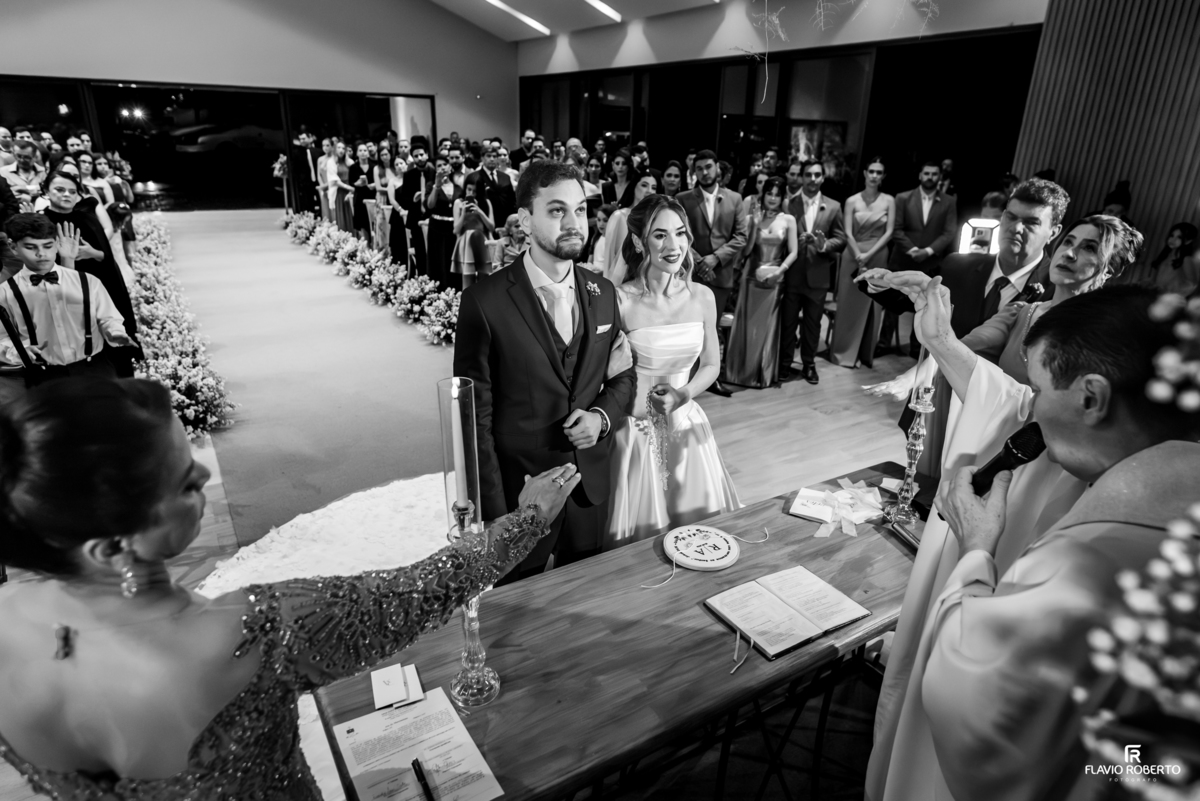 Noivos recebem bênção durante cerimônia católica de casamento com convidados ao fundo no Recanto do Bosque.