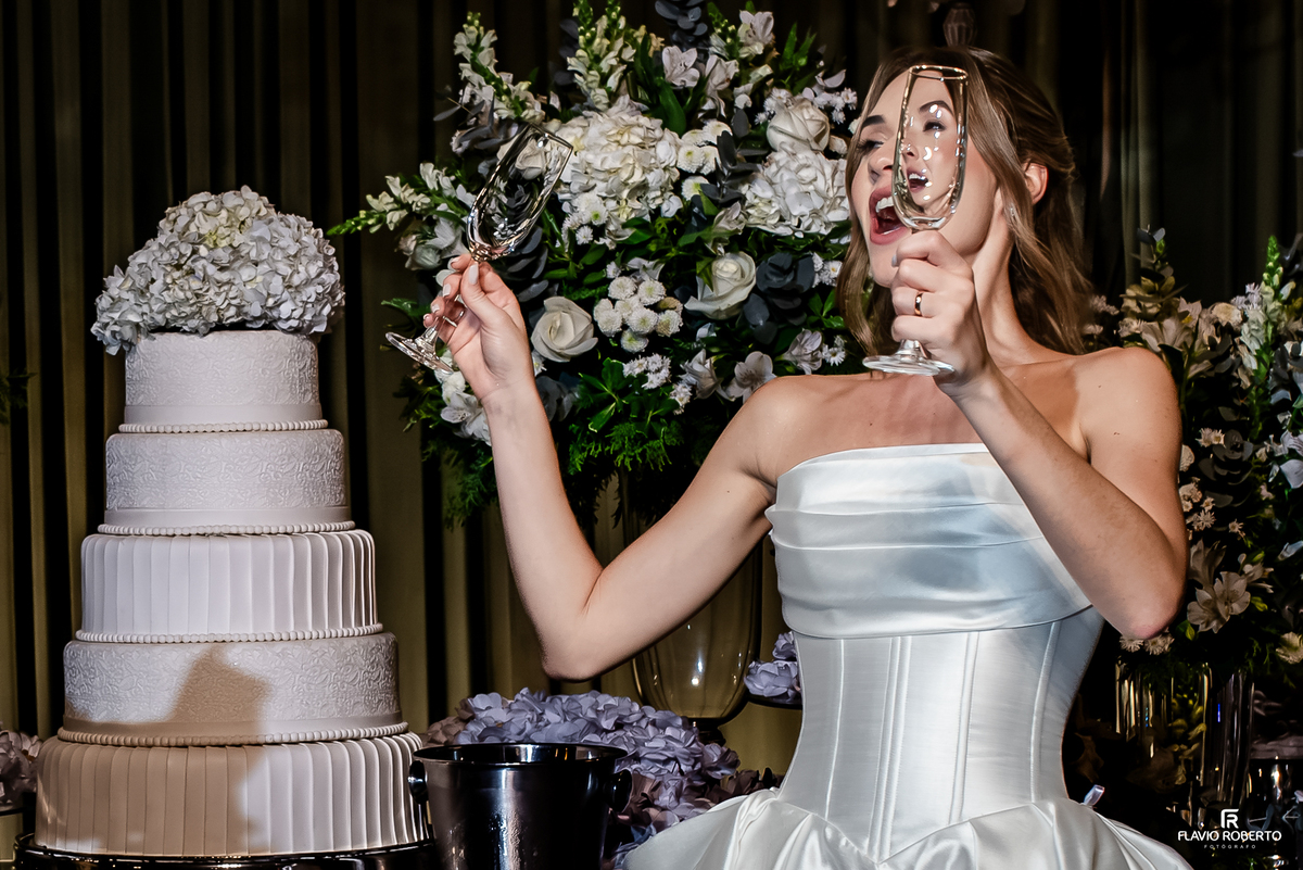Noiva segura duas taças em momento divertido ao lado do bolo durante festa de casamento em Guaratinguetá.