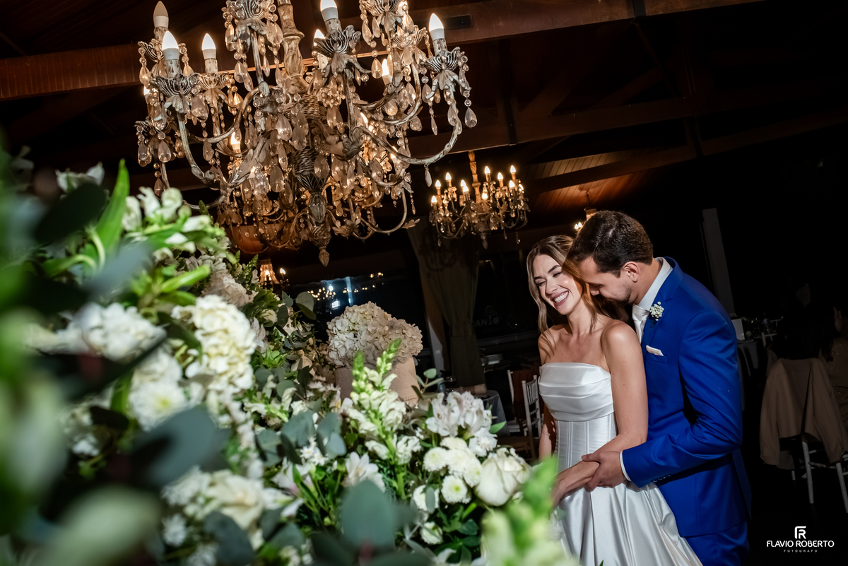 Noivos abraçados e sorrindo junto à decoração floral da recepção de casamento no Recanto do Bosque.