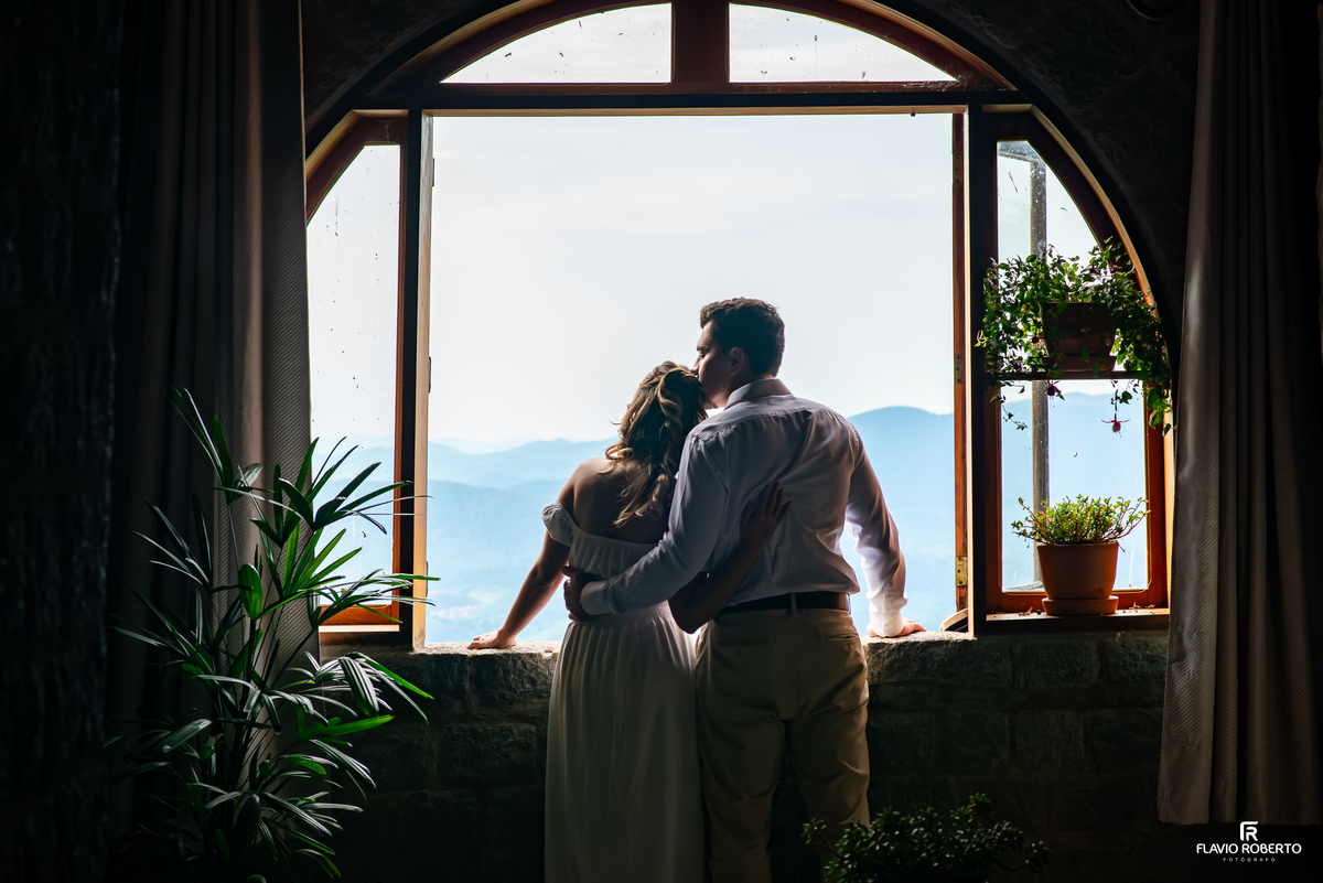 Ivanna e Luis Gustavo abraçados diante da janela com vista para as montanhas durante pré-wedding no Castelo de Pedra em Itamonte MG.