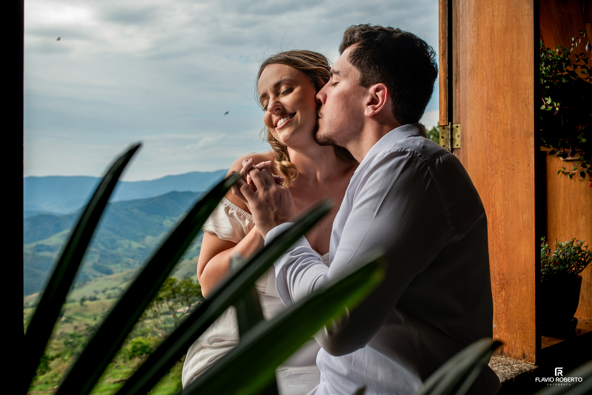 Ivanna recebe beijo de Luis Gustavo junto à janela com vista para as montanhas em ensaio de casal em Itamonte MG.