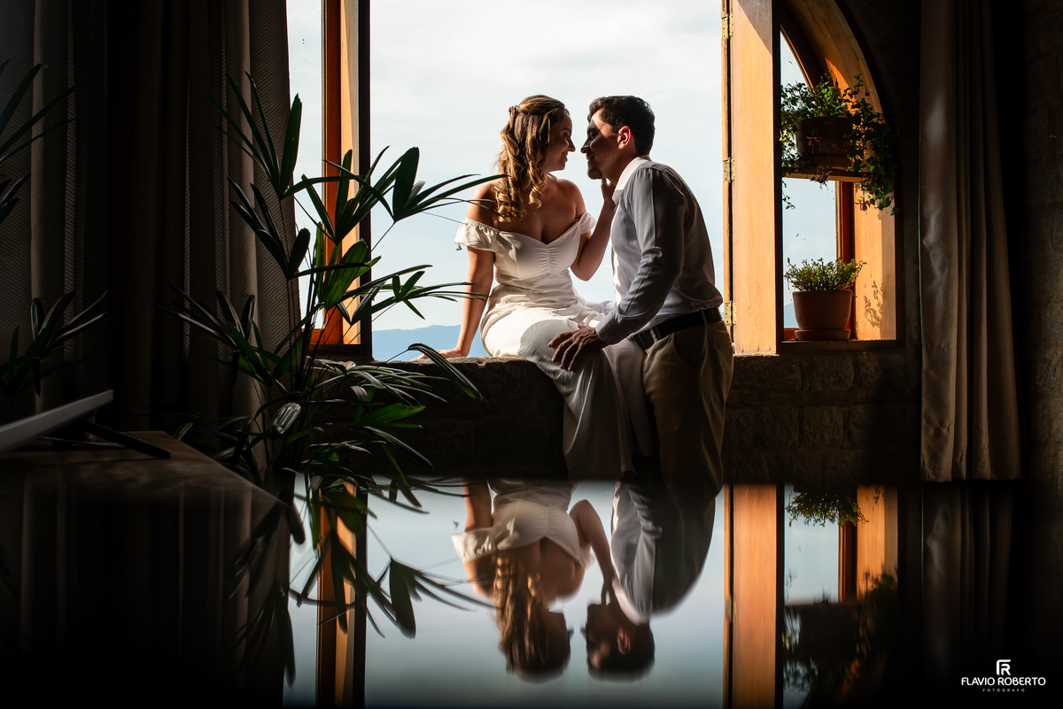 Ensaio pré-wedding de Ivanna e Luis Gustavo junto à janela do Castelo de Pedra, com reflexo e paisagem das montanhas de Itamonte.