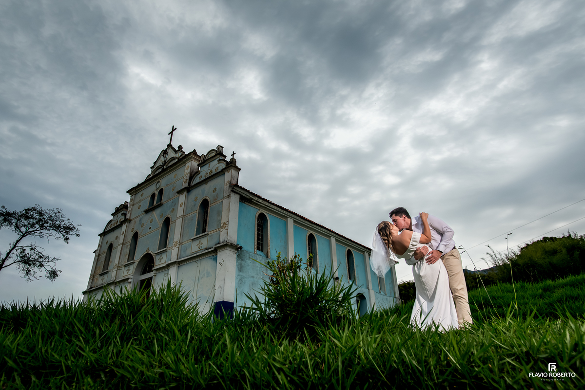 Ivanna e Luis Gustavo em fotografia ampla diante da Igreja Nossa Senhora da Conceição sob céu dramático em Itamonte MG.