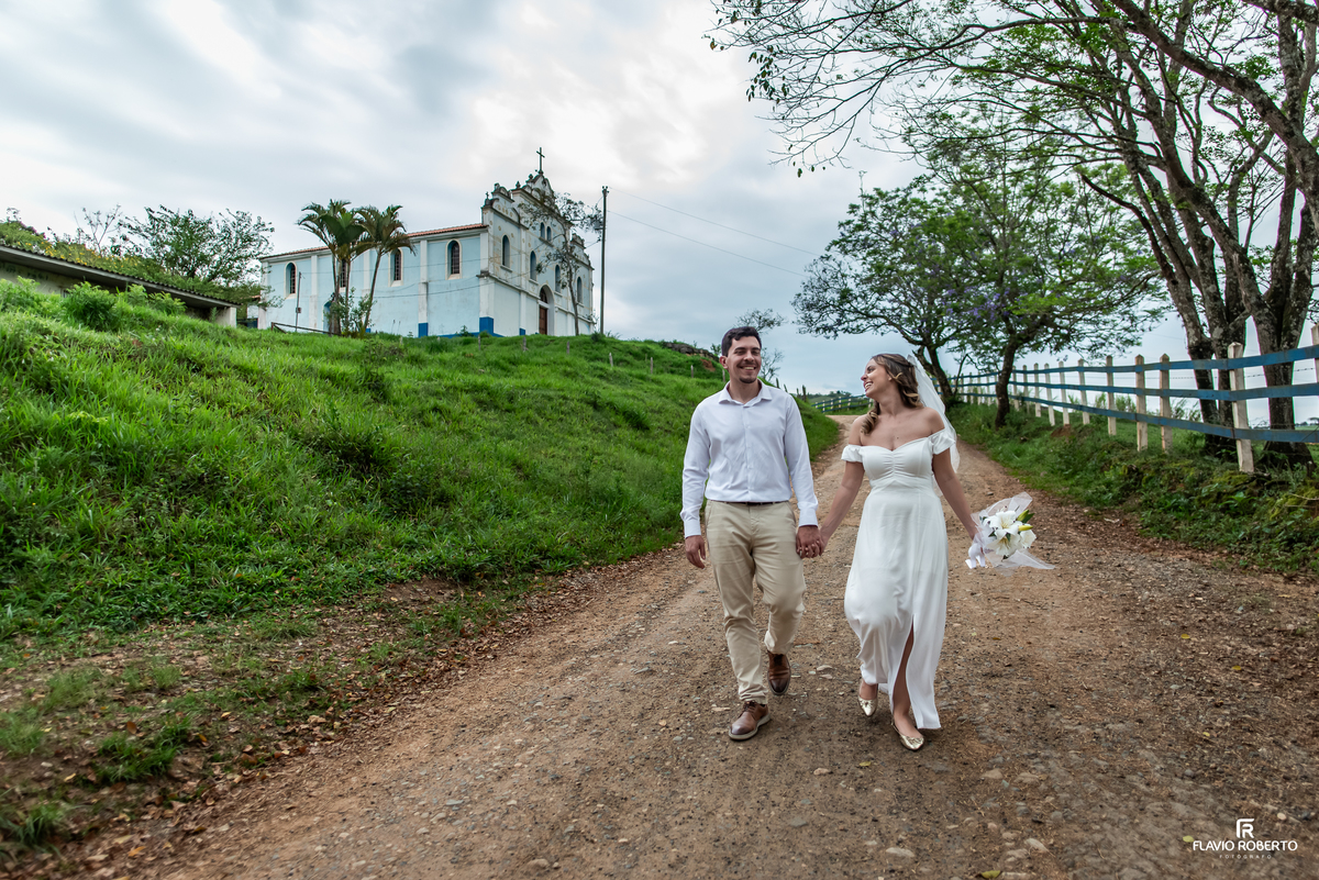 Casal caminhando de mãos dadas por estrada rural com a Igreja Nossa Senhora da Conceição ao fundo em Itamonte, Minas Gerais.
