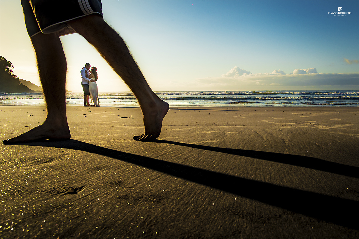 noivos abraçados no meio das pernas de um turista caminhado na praia