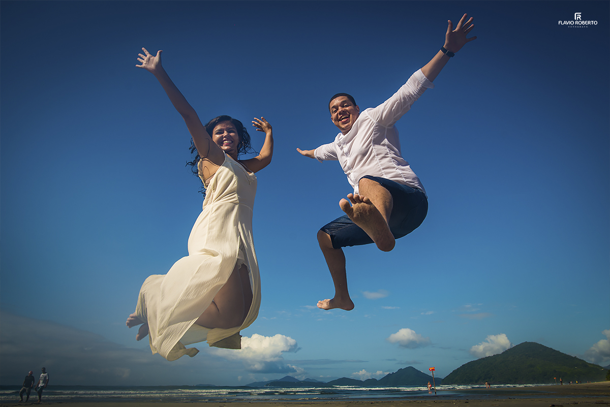 Casal de Noivos saltando bem alto na praia, com um céu azul de fundo