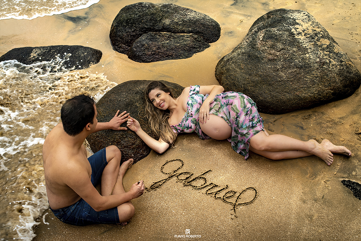 revelando o nome do filho durante o ensaio de gestante na praia. casal deitado na praia escrevendo na areia no Ensaio de Gestante em Ubatuba