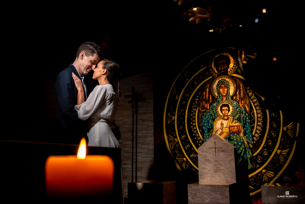 Noivos abraçados atrás de uma vela do lado do ícone da Sagrada Família na Paróquia Sao Luiz Gonzaga em SP