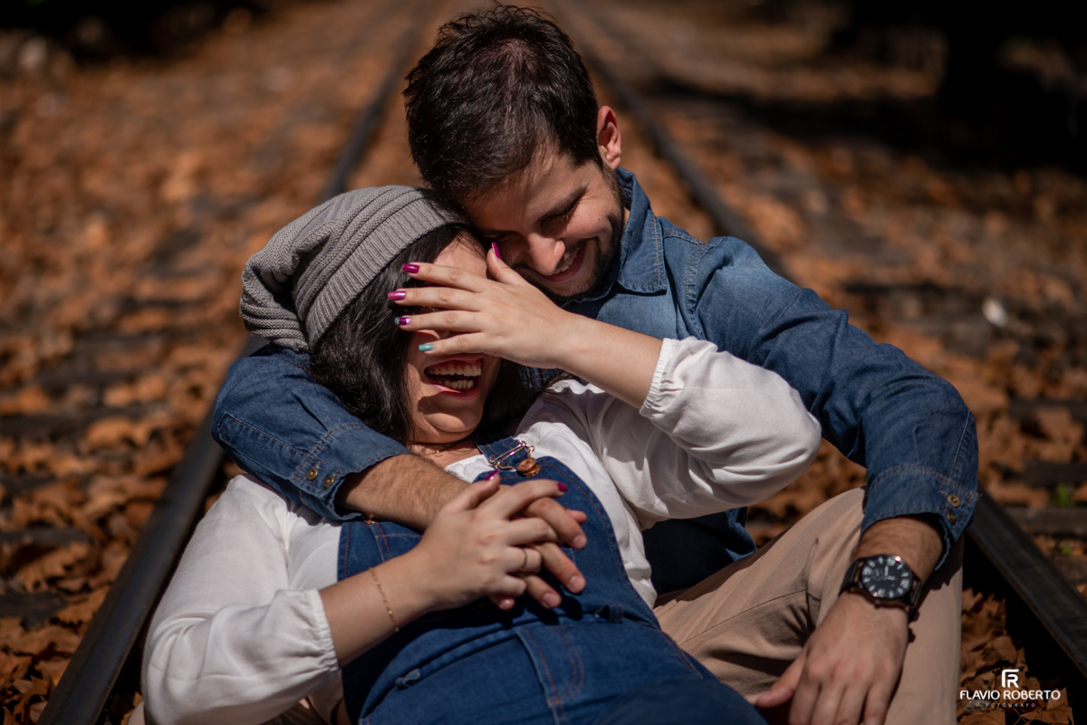 Casal abraçado e rindo sentados na linha de trem entre as folhas secas do outono de Campos do Jordão no Ensaio Pré Wedding.