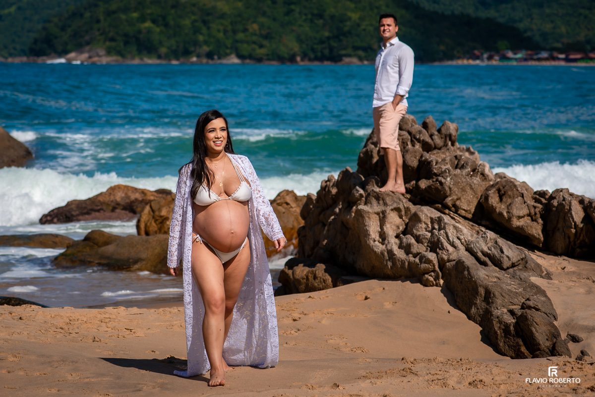 Gestante caminhando na praia em Trindade, sendo observada por seu esposo em Ensaio de Gestante em Paraty-RJ.