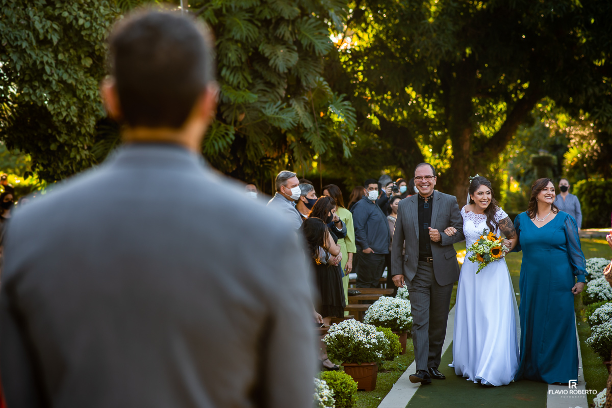 Noiva entrando com seus pais, sendo observada pelo noivo  em Casamento realizado no Sítio dos Ipês em Taubaté