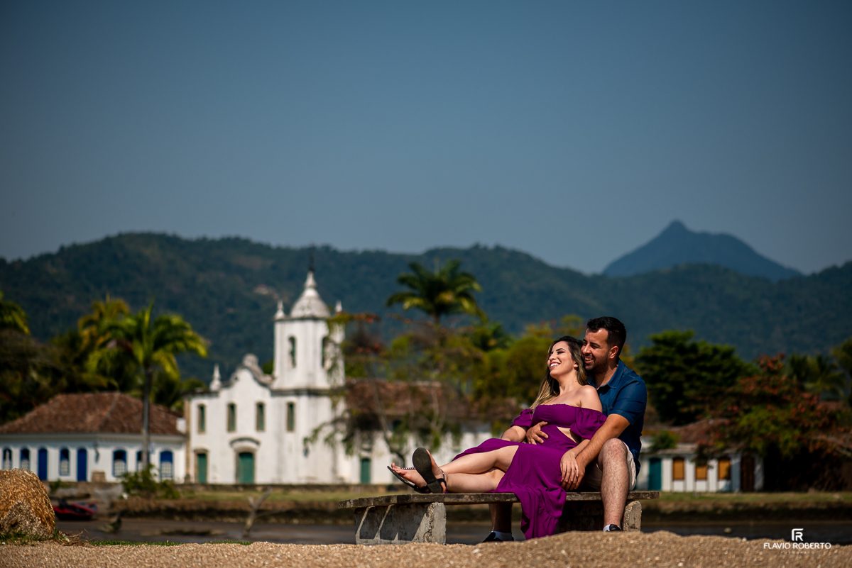 Noivos sentados em um banquinho do lado da praia namorando com uma igrejinha de fundo, durante Ensaio Pre Wedding no centro historico de Paraty-RJ
