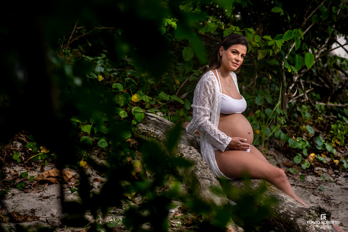 Gestante sentada em um troco entre folhagens verdes durante ensaio de gestante na praia de Ubatuba 