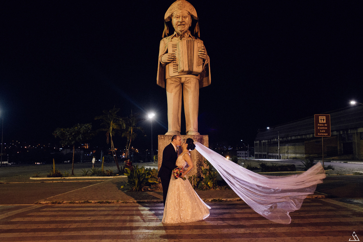 Jessica Mendes fotografia de casamento bárbara e itaguaraci. Fotógrafo de casamento em Pernambuco. Caruaru Recife. noivos na estátua de luiz gonzaga no pátio do forró em Caruaru - PE. Foto do véu da noiva voando.