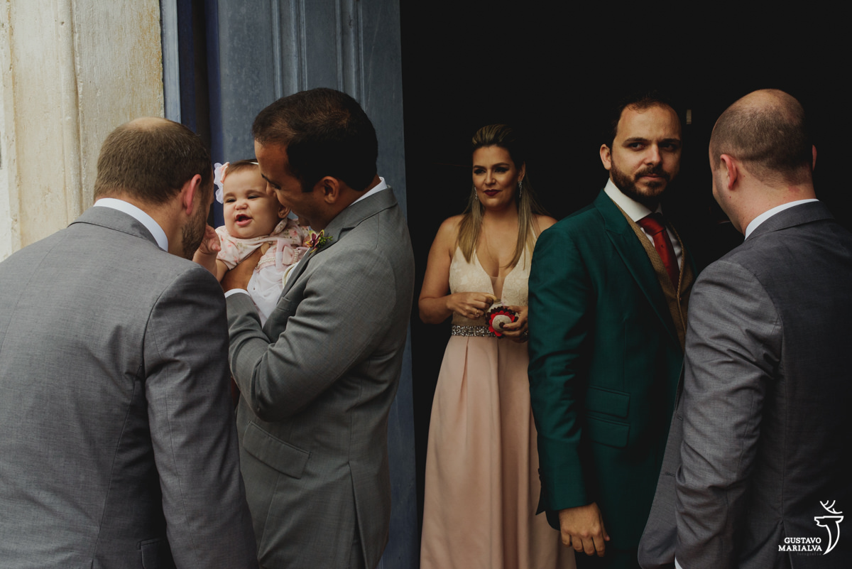 homem segura bebe no colo enquanto amigo faz ela sorrir e noivo observa tenso antes da cerimônia do casamento