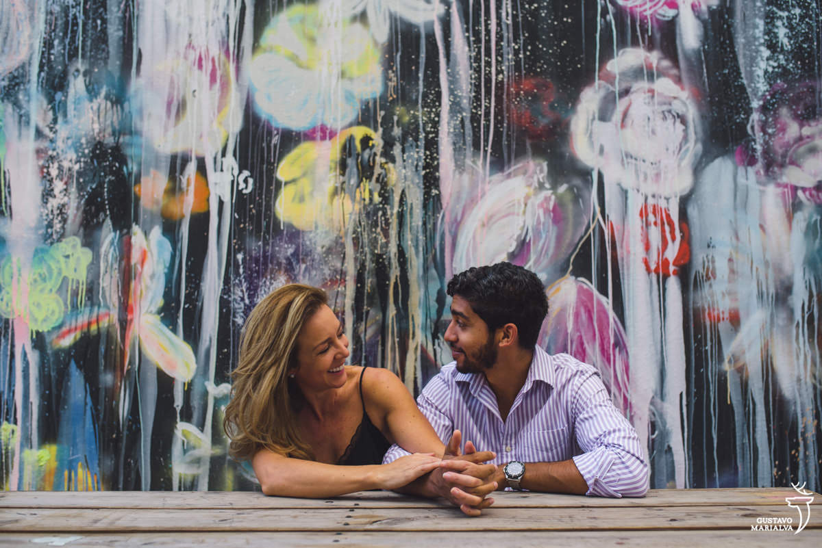 casal sentados em banco dá as mãos e se olham sorrindo com parede grafitada com flores ao fundo