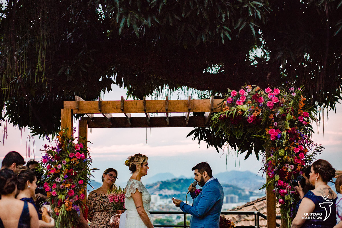 noivo lê os votos e é observado por noiva com gazebo e madeira e flores rosas