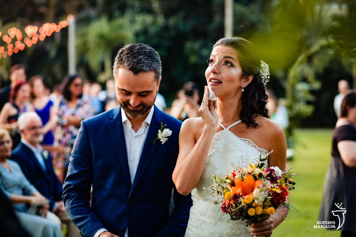 noiva segurando buquê e chorando olha para cima enquanto é observada pelo noivo que usa terno azul na cerimônia de casamento
