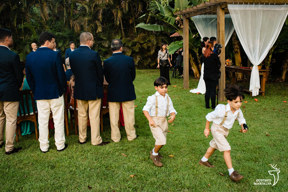 meninos brincam de correr usando camisa branca e suspensório bege enquanto convidados observam a cerimônia de casamento