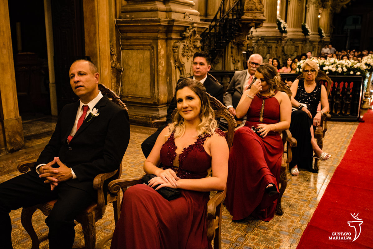 padrinhos usando terno preto e gravata vinho observam a cerimônia de casamento enquanto as madrinhas usando vestido vinho choram
