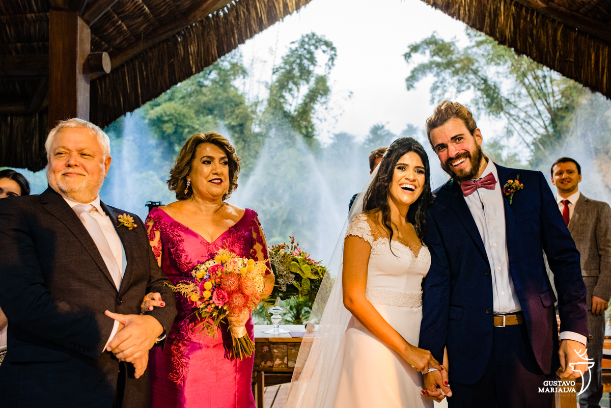 noivos observam entrada dos pajens abraçados e emocionados enquanto são observados pela mãe que segura o buquê de noiva