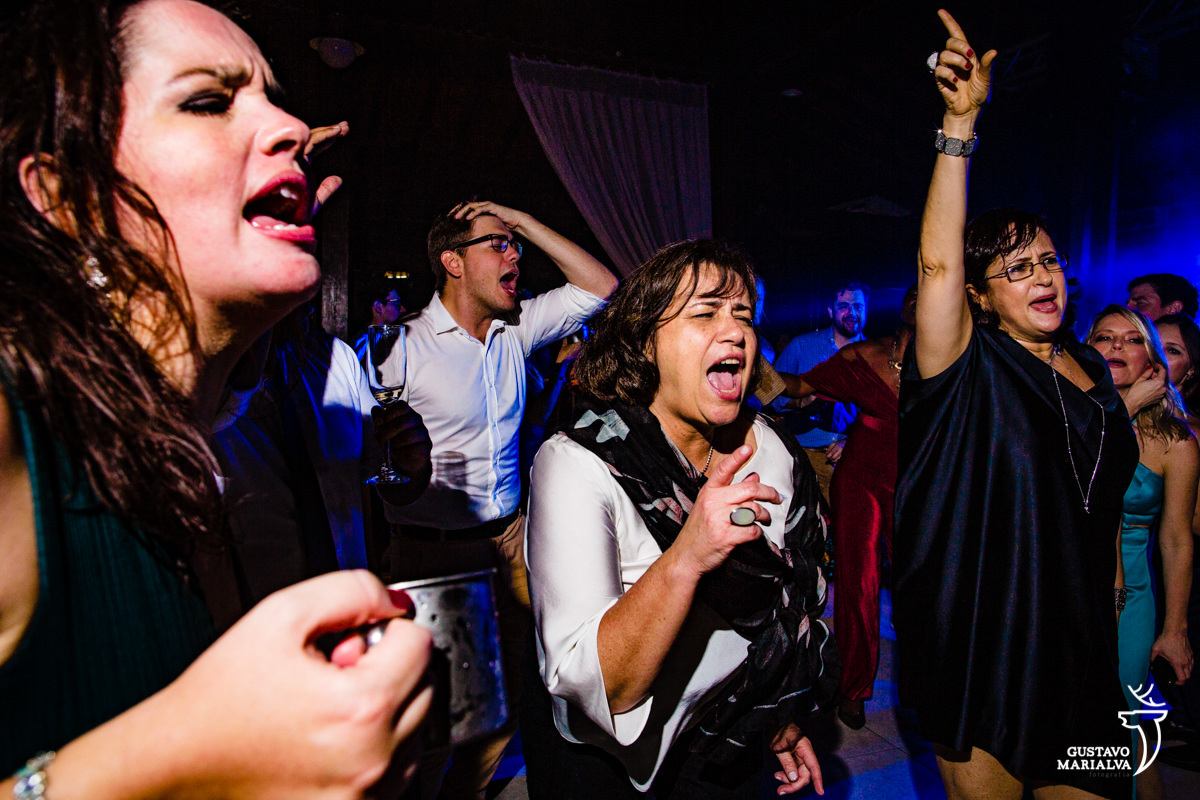mulher usando roupa branca dança enquanto amiga faz pose com as mãos para cima e homem dança com as mãos na cabeça