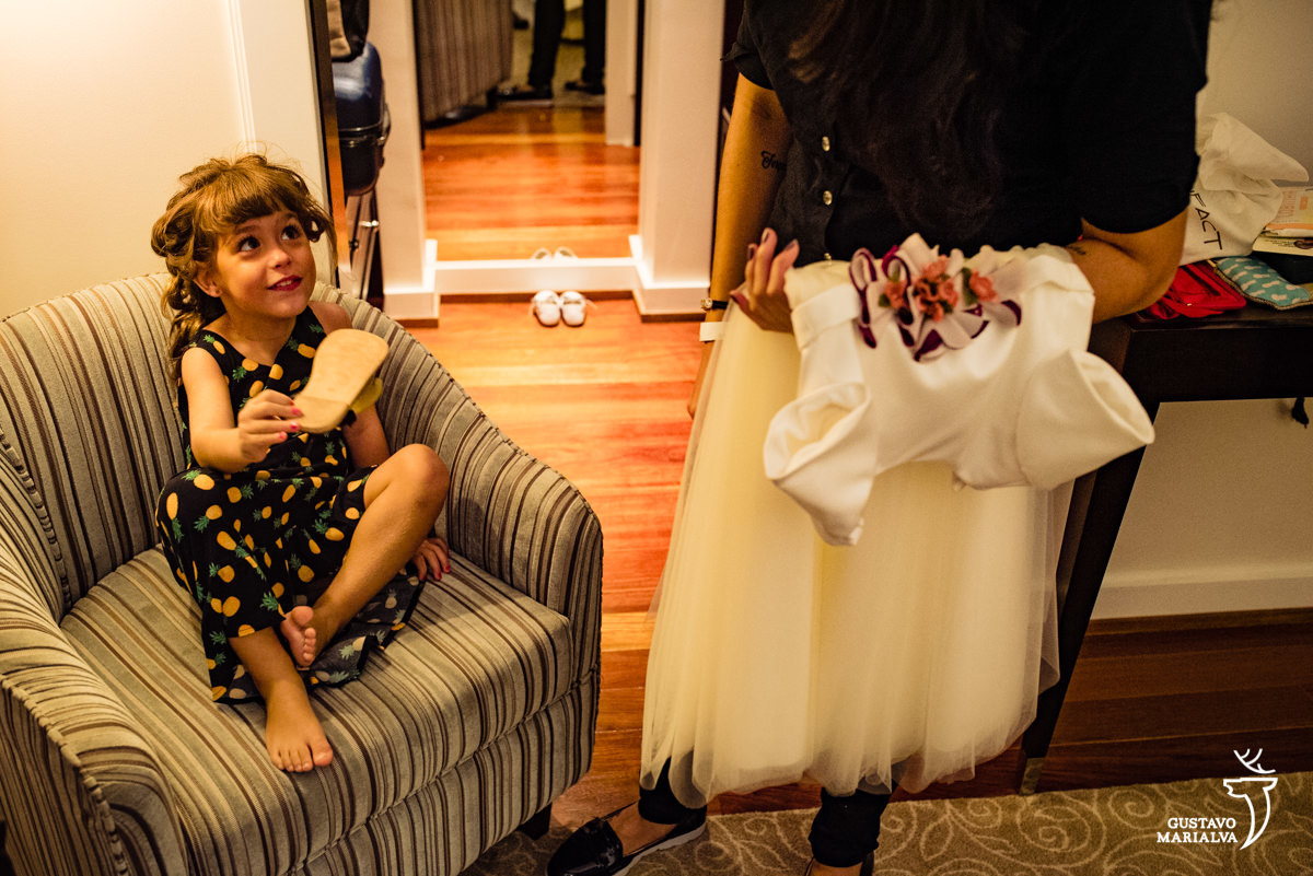 menina com sandália na mão faz careta para mulher que segura vestido da daminha