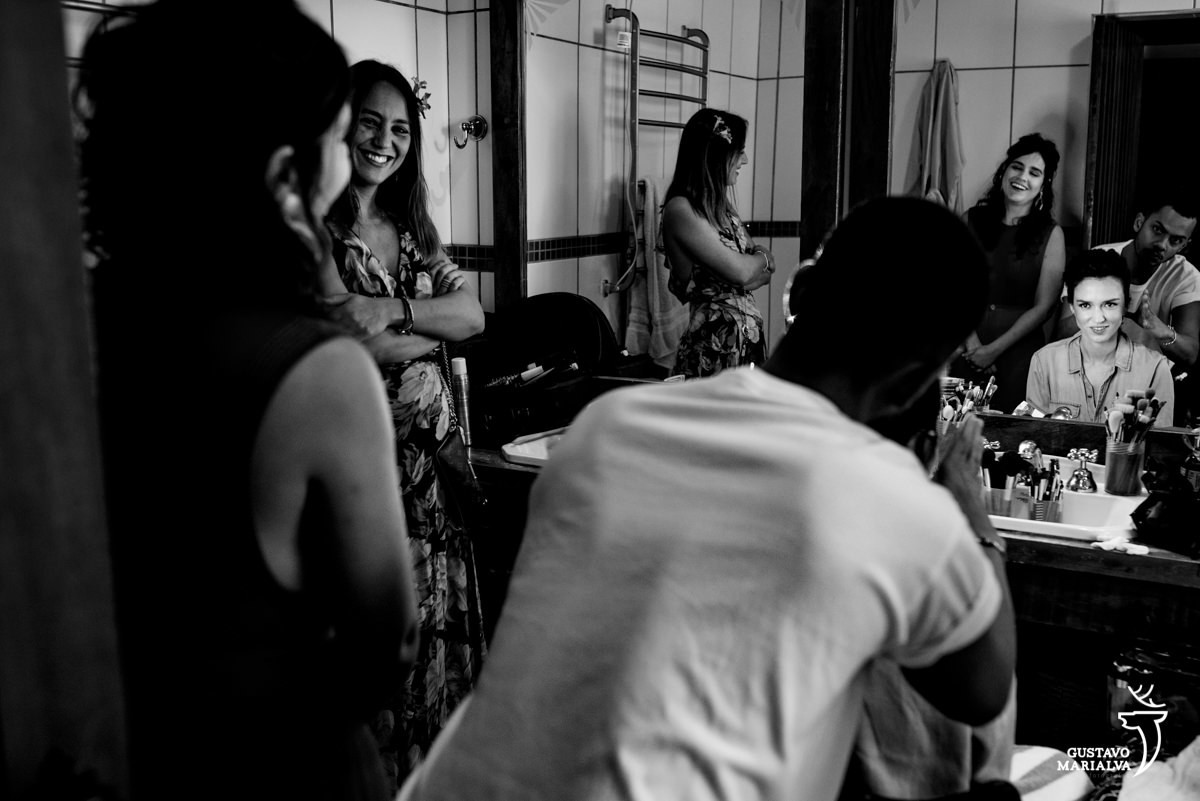 noiva se olha no espelho enquanto arruma o cabelo e amigas sorriem dentro do banheiro