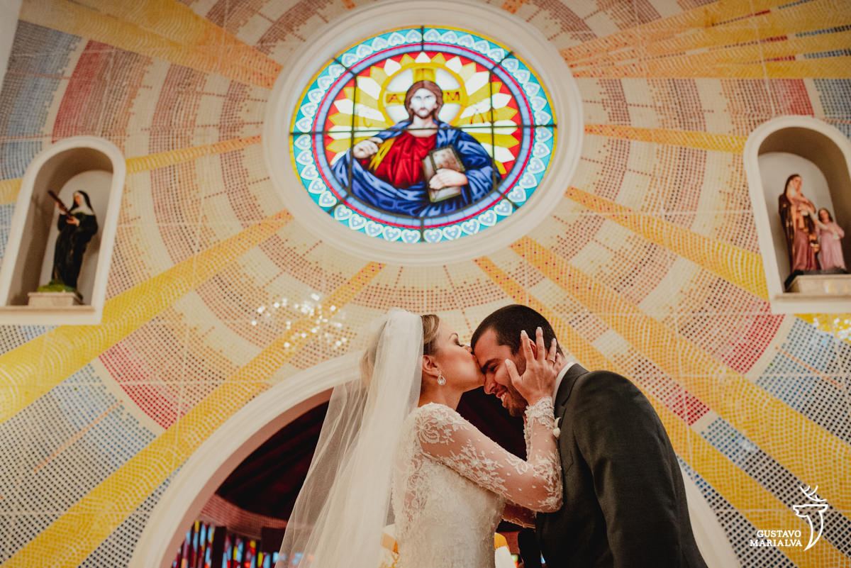 noiva beija a testa do noivo com imagem de jesus ao fundo durante a cerimônia de casamento