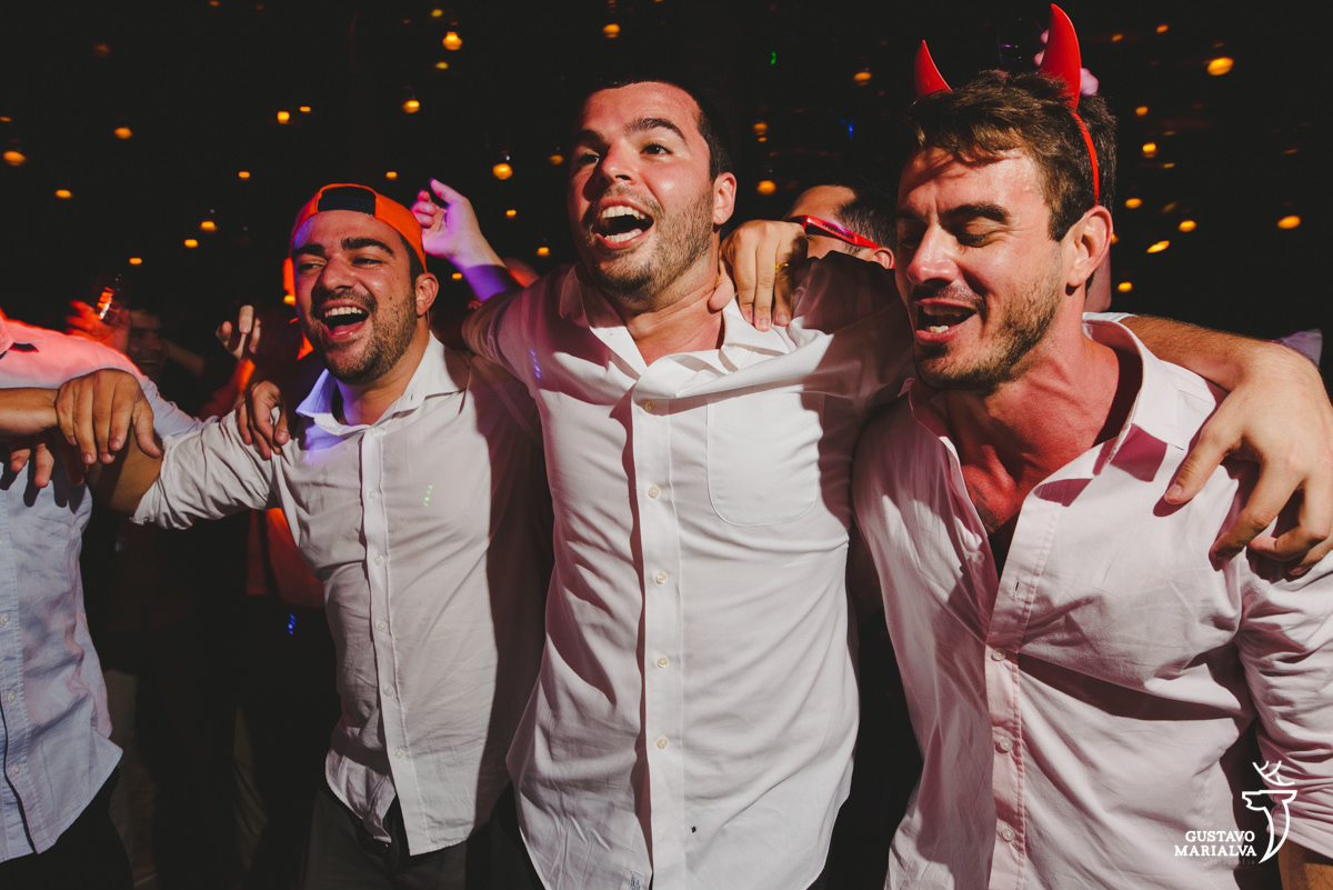 noivo dança abraçado a amigo que usa boné laranja e outro que usa arco de diabinho