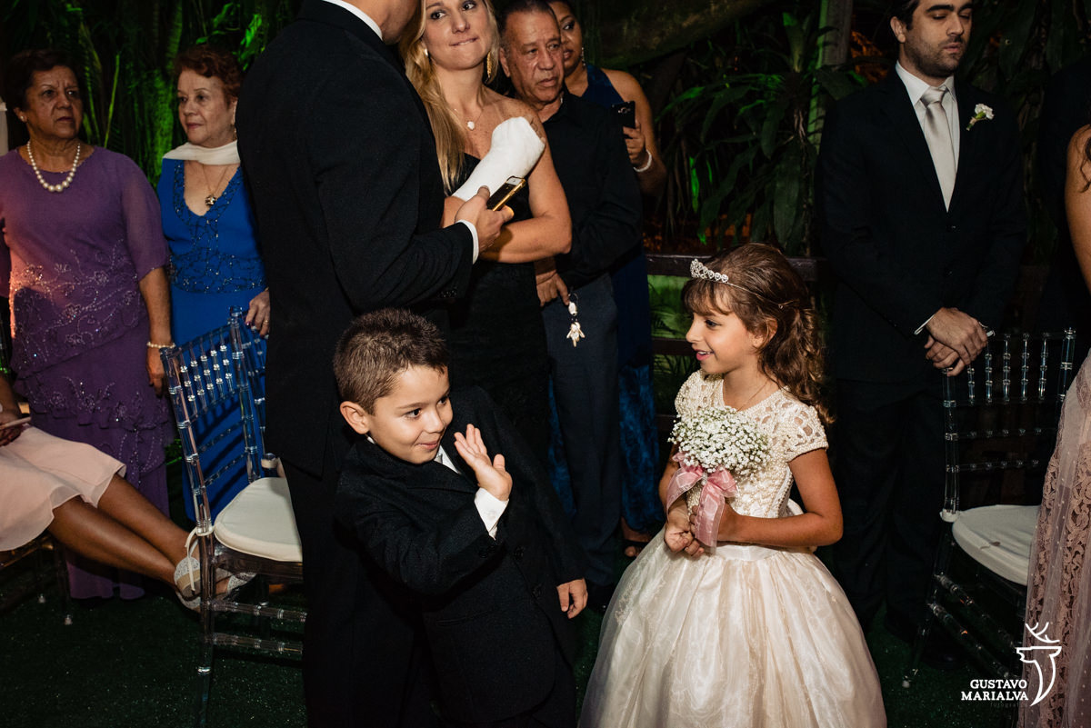 pajem usando terno preto acena para madrinha que usa vestido branco e segura buque de rosas brancas e são observados por mulher com vestido azul