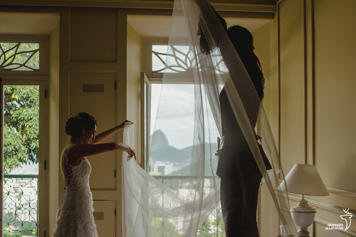 noiva usando vestido de noiva ajeita o véu na cama enquanto é ajudada por maquiadora